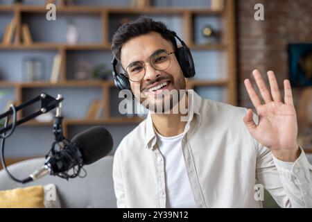 Fröhlicher junger Mann mit Brille und Kopfhörern, der vor der Kamera winkt, während er in einem modernen Studio Podcasts aufnimmt. Er sitzt in lässiger Kleidung mit Mikrofon und drückt Freundlichkeit und Begeisterung aus. Stockfoto