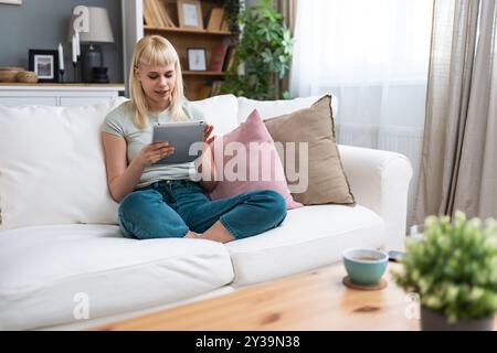 Glückliches Teenager-Mädchen mit Pad-Computer-Gadget mit digitaler Tablet-Technologie, das zu Hause auf der Couch sitzt. Lächelnde junge Frau, die Apps nutzt, einkaufen, online Stockfoto