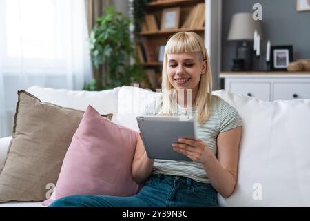 Glückliches Teenager-Mädchen mit Pad-Computer-Gadget mit digitaler Tablet-Technologie, das zu Hause auf der Couch sitzt. Lächelnde junge Frau, die Apps nutzt, einkaufen, online Stockfoto