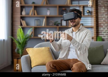 Junger Mann, der mit dem VR-Headset in die virtuelle Realität eingetaucht ist, auf dem Sofa saß und mit der virtuellen Umgebung interagierte. Begeistert lächeln und hochmoderne Technologie erleben Stockfoto