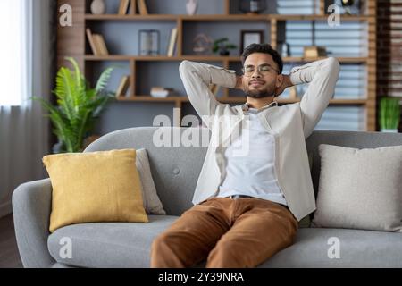 Junger Mann, der sich auf dem Sofa auszieht, die Augen geschlossen, lässige Kleidung trägt. Entspannende Innenräume, friedliche und zufriedene Atmosphäre. Gemütliche Wohnzimmerumgebung mit bequemen Kissen. Stockfoto