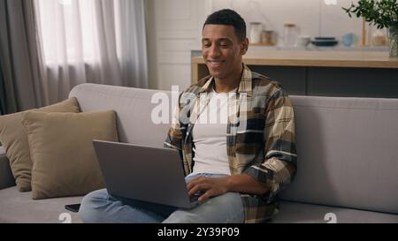 Glücklicher lächelnder afroamerikanischer ethnischer Mann, der zu Hause arbeitet entfernte Online-Arbeit studiert auf Sofa Guy Laptop mit Computer auf Couch männlich mit pc Stockfoto