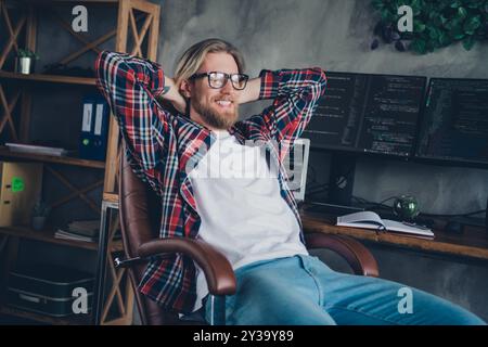 Foto eines fröhlichen, selbstbewussten, gutaussehenden, bärtigen Programmierers, der auf einem Sessel sitzt und mit Computermonitoren im Spielzimmer sitzt Stockfoto