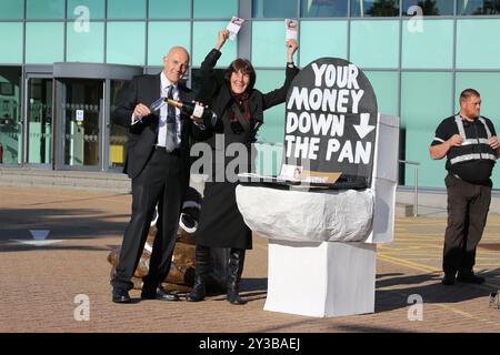 Huntingdon, England, Großbritannien. September 2024. Als wohlhabende Aktionäre gekleidete Demonstranten winken Geld neben einem riesigen Modellloo vor den Büros von Anglia Water. Demonstranten von Extinction Rebellion in Gefahrenanzügen mit Theaterauflagen demonstrieren vor den Büros von Anglian Water. Sie protestieren gegen die Bilanz der Wassergesellschaft bei der Ableitung von Rohabwasser in Meere und Flüsse in ihrer Region. Im Jahr 2023 gab es in der Wasseraufbereitungsanlage in Cambridge 74 ausgelaufene Flüssigkeiten, die über 1476 Stunden dauerten. Sie argumentieren, dass die Kosten für die Verhinderung dieses Problems unter den 78 Milliarden Pfund liegen, die Â an die Aktionäre ausgezahlt wurden. (Kreditbild: Stockfoto