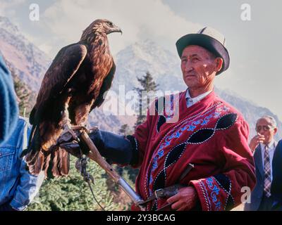 Ein traditioneller kirgisischer Adlerjäger, der vor einer bergigen Kulisse steht, in lebendiger, traditioneller Kleidung gekleidet und einen majestätischen Adler hält. Stockfoto