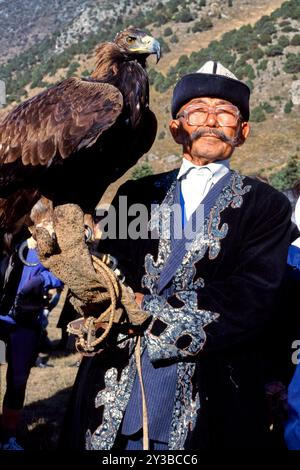 Älterer kirgisischer Mann, der das kulturelle Erbe mit einem majestätischen Adler präsentiert. Traditionelle Kleidung und die Bergkulisse betonen die Essenz des Adlers Stockfoto