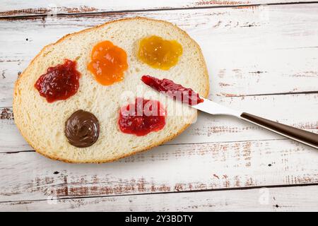 Scheibe Weißbrot mit verschiedenen Marmeladenaufstrichen und Messer. Blick von oben auf Holztisch. Stockfoto