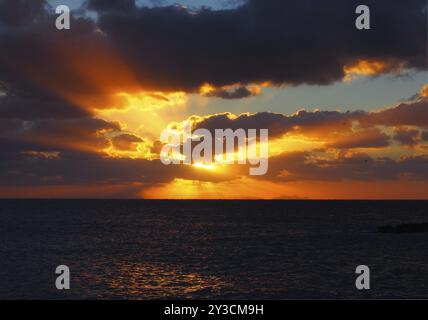 Sonnenuntergang über einem ruhigen dunklen Meer mit orangen Strahlen, die durch dunkle dramatische Abendwolken leuchten Stockfoto