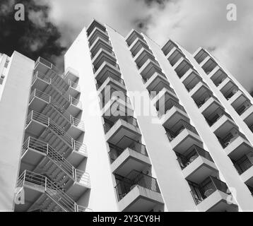 Monochromer, abgewinkelter Detailblick auf ein altes weißes Betonwohnhaus aus den 1960er Jahren mit Stufen und Balkonen gegen Himmel und Wolken Stockfoto