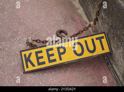 Ein fluoreszierendes gelbes Bleib-Schild aus Metall auf einem Betonboden mit rostiger Kette, die es mit einem Eisenhaken an einer Wand befestigt Stockfoto