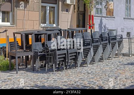 Gestapelte schwarze Rattan-Stühle und -Tische vor dem Coffee Shop in der Kopfsteinpflasterstraße Stockfoto