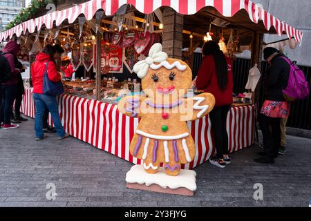 Eine Lebkuchenfrau vor einem Weihnachtsmarkt, voll mit Leuten, die in mehr London Place, Großbritannien, festliche Leckereien und Süßigkeiten kaufen. Stockfoto