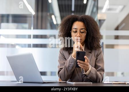Nahaufnahme einer jungen afroamerikanischen Frau in einem Anzug, die am Schreibtisch im Büro sitzt, den Kopf in der Hand hält und nachdenklich auf den Bildschirm ihres Mobiltelefons blickt. Stockfoto