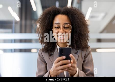 Foto einer lächelnden afroamerikanischen Frau mit lockigen Haaren in Business-Anzug und Brille, die im Büro sitzt und das Handy benutzt. Stockfoto