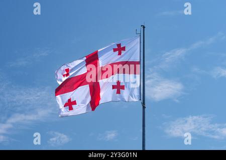 Die georgische Flagge winkt gegen einen klaren blauen Himmel und symbolisiert Stolz und nationale Identität. Die Flagge von Georgia flattert an einem hellen, sonnigen Tag auf einem Fahnenmast. Stockfoto