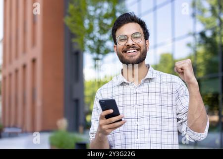 Nahporträt eines jungen muslimischen Mannes, der vor einem Gebäude steht, ein Telefon hält, eine Handbewegung macht und in die Kamera lächelt. Stockfoto