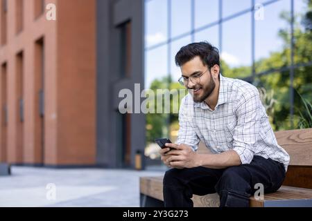 Ein lächelnder junger Indianer mit Brille und Hemd sitzt auf einer Bank in der Nähe eines Gebäudes und benutzt ein Handy. Stockfoto