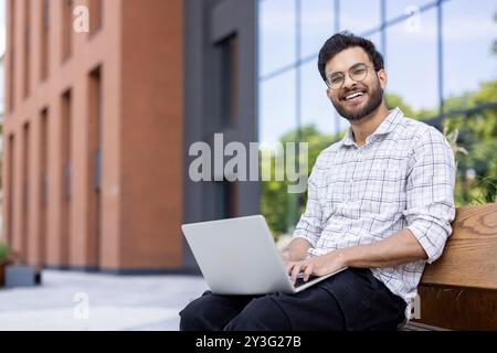 Porträt eines jungen muslimischen Mannes mit Brille, der auf einer Bank in der Nähe eines Bürogebäudes sitzt, einen Laptop auf seinem Schoß hält und in die Kamera lächelt. Stockfoto