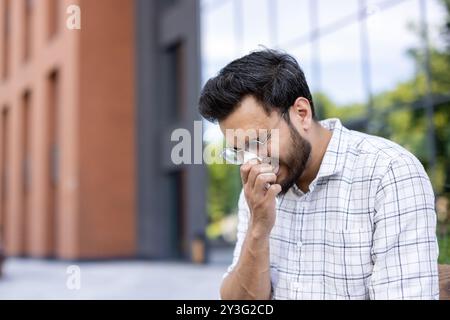 Nahaufnahme eines jungen kranken Indianers, der draußen auf der Straße in der Nähe eines Bürozentrums sitzt und sich mit einem Taschentuch die Nase abwischt, niest und seinen Mund mit der Hand bedeckt. Stockfoto