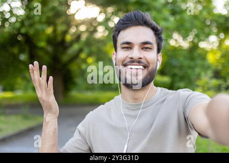 Junger Mann lächelt beim Selfie im Park mit Smartphone und Ohrhörern. Fängt Glück, Verbindung und entspannte Atmosphäre im Freien in der Freizeit ein. Stockfoto
