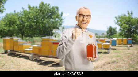 Ein älterer Mann, der auf einer Bienenfarm einen Holzlöffel und ein Honigglas hält Stockfoto