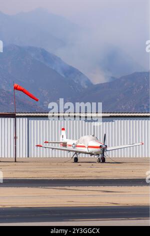 Ein privates Tiefflügelflugzeug ist in der Nähe eines Windindikators fixiert, was auf mäßige Windaktivität hinweist. Waldbrandrauch ist auf einem nahegelegenen Berg im Hintergrund zu sehen. Stockfoto
