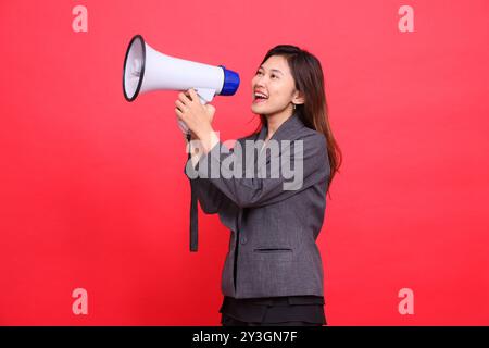 Fröhlicher weiblicher Ausdruck im asiatischen Büro, schräg oben rechts, schreiend mit einem Megaphon, offen mit grauer Jacke und Rock auf rotem Hintergrund. f Stockfoto