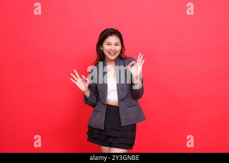 Fröhlicher Ausdruck einer indonesischen Bürofrau mit beiden Händen, die Gunst zeigen (okay, gut), tragen eine graue Jacke und einen Minirock auf rotem Hintergrund. Für Stockfoto