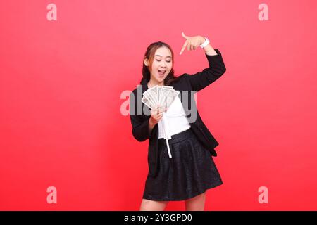 Geste einer jungen asiatischen Bürofrau, die Geld ansieht, glücklich in einem schwarzen formalen Anzug, der einen Dollarschein hält und von oben auf ihn zeigt, für Dekor Stockfoto