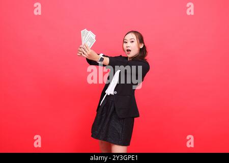 Geste der jungen indonesischen Bürofrau glücklich überrascht über die Kamera, die einen schwarzen formellen Anzug trägt, beide Hände halten Dollarscheine vor dem R Stockfoto