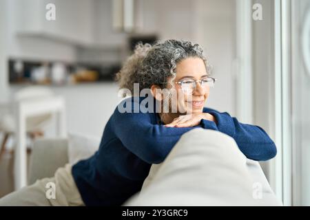 Glückliche reife Frau sitzt auf dem Sofa im Wohnzimmer zu Hause und blickt durch das Fenster. Stockfoto