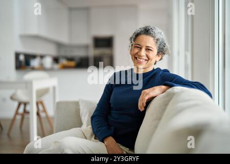 Glückliche, reife Frau, die zu Hause auf der Couch sitzt und in die Kamera schaut, Porträt. Stockfoto