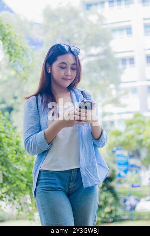 Schöne asiatische Frau lächelt mit Smartphone, während sie Ohrhörer im Campus Park an sonnigen Sommertagen trug. Studentinnen für Bildung, Promotion A Stockfoto