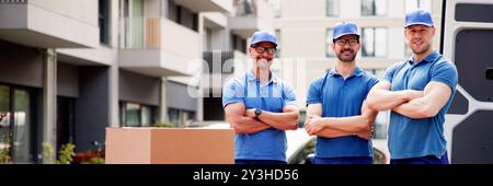 Junge und glückliche männliche Umzugswagen in Schutzanzügen, die vor einem Lieferwagen stehen und Kisten für ein umziehendes Serviceunternehmen liefern. Stockfoto