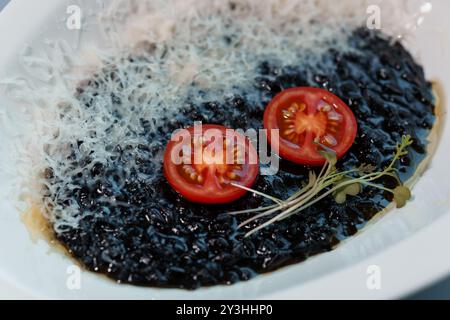 Schwarzer Gourmet-Risotto mit geriebenem Käse und frischen Kirschtomaten garniert mit Microgreens auf einer eleganten weißen Platte. Stockfoto