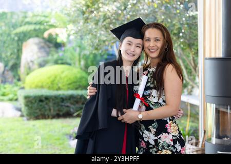 Im Garten, Abitur, asiatische Tochter in Mütze und Kleid umarmt lächelnde Mutter draußen Stockfoto