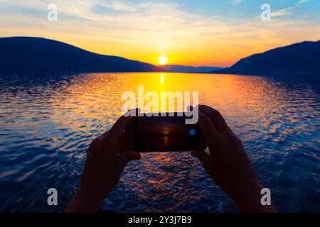 Aufnahmen eines atemberaubenden Sonnenuntergangs mit Smartphone, Silhouetten vor dem pulsierenden Himmel, Bergen und Meer. Die Person fotografiert den Sonnenuntergang über einem Stockfoto