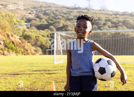 Draußen, lächelnder afroamerikanischer Junge, der Fußball auf dem Schulfeld hält Stockfoto