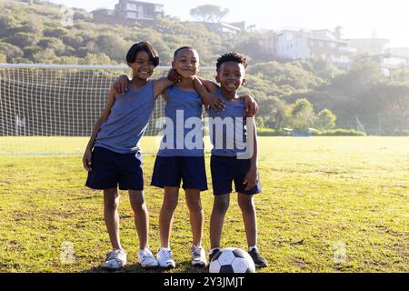 Draußen, Fußball spielen, drei multirassische Jungs in Sportbekleidung lächeln auf dem Schulfeld Stockfoto