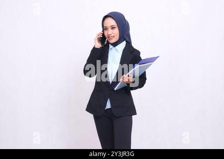 Exklusive indonesische Bürofrau mit Hijab steht lächelnd und ruft Leute an und trägt einen Stift, ein Klemmbrett und ein Notizbuch. Schöne muslimische Frau in Stockfoto