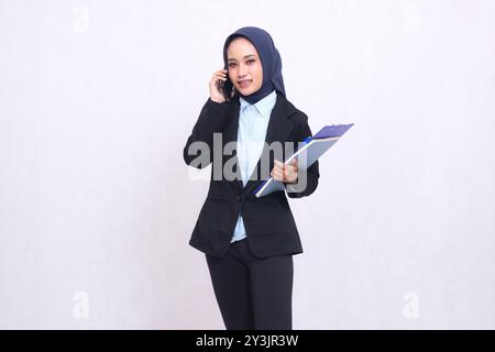 Eine exklusive asiatische Bürofrau mit Hijab steht lächelnd auf die Telefonkamera und trägt einen Stift, ein Klemmbrett und ein Notebook. Wunderschöne muslimische Frau Stockfoto