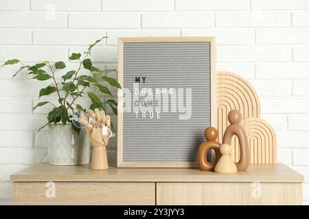 Brieftafel mit dem Satz My Dreams Come True, Pflanzen- und Dekorationselemente auf einem hölzernen Beistelltisch in der Nähe der weißen Ziegelwand Stockfoto