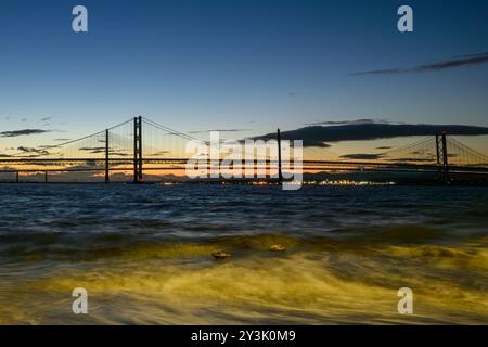 Ein Blick auf die Forth Rail Bridge vor Einem Sonnenuntergang mit der Queensferry Crossing Bridge im Hintergrund und River Forth Waves im Vordergrund Stockfoto