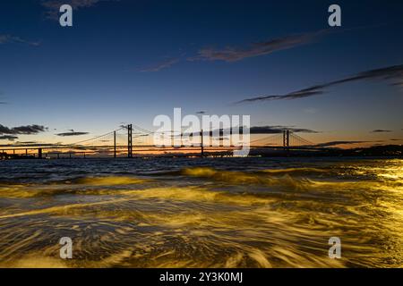 Ein Blick auf die Forth Rail Bridge vor Einem Sonnenuntergang mit der Queensferry Crossing Bridge im Hintergrund und River Forth Waves im Vordergrund Stockfoto