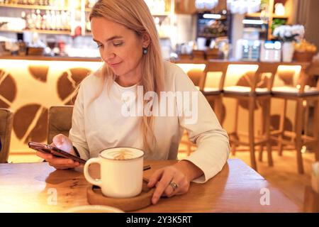 Eine junge Frau hält ein Telefon und trinkt Kaffee in einem Café. Eine hübsche Blondine plaudert und isst zu Mittag. Ein charmantes Mädchen kauft online oder arbeitet Stockfoto