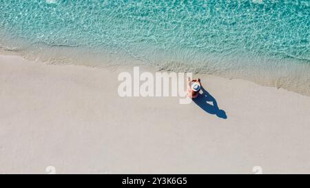 Ein Blick aus der Luft auf einen jungen Mann mit weißem Hut, der am Meer sitzt. Ein Mann genießt die Meereslandschaft. Ein Mann entspannt sich an einem sauberen Sandstrand. Urlaub Stockfoto