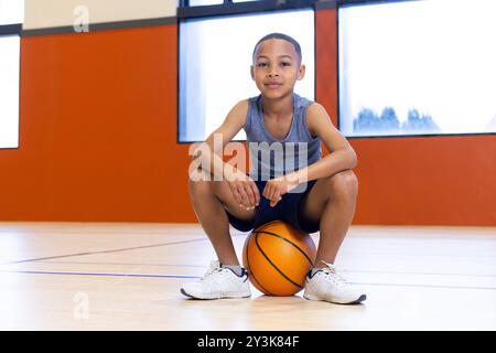 In der Turnhalle sitzt der Junge auf Basketball, sieht selbstbewusst und entspannt aus Stockfoto