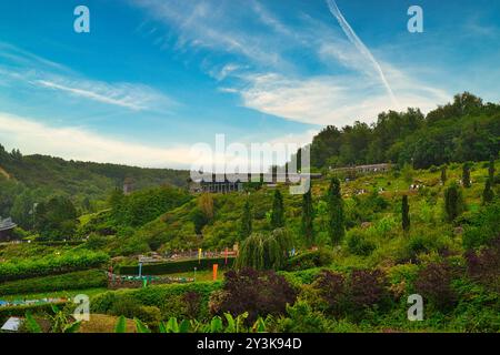 Ein malerischer Blick auf einen üppig grünen Hügel mit einem modernen Gebäude inmitten von Bäumen. Der Himmel ist hellblau mit schimmernden Wolken, und farbenfrohe Gärten sind es Stockfoto