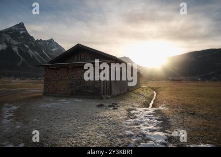 Holzscheune mit einem Haufen Holz auf einer Wiese mit Schnee und getrocknetem Gras, in den Alpen, bei Sonnenuntergang, im Winter Stockfoto