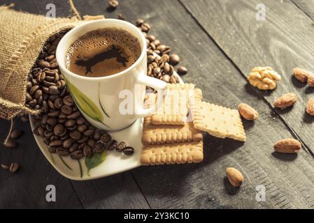 Kaffee Tasse auf einem Teller und Kaffeebohnen Ausgießen aus Sackleinen Sack, Kekse, Walnüsse und Mandeln auf einem Holztisch Stockfoto